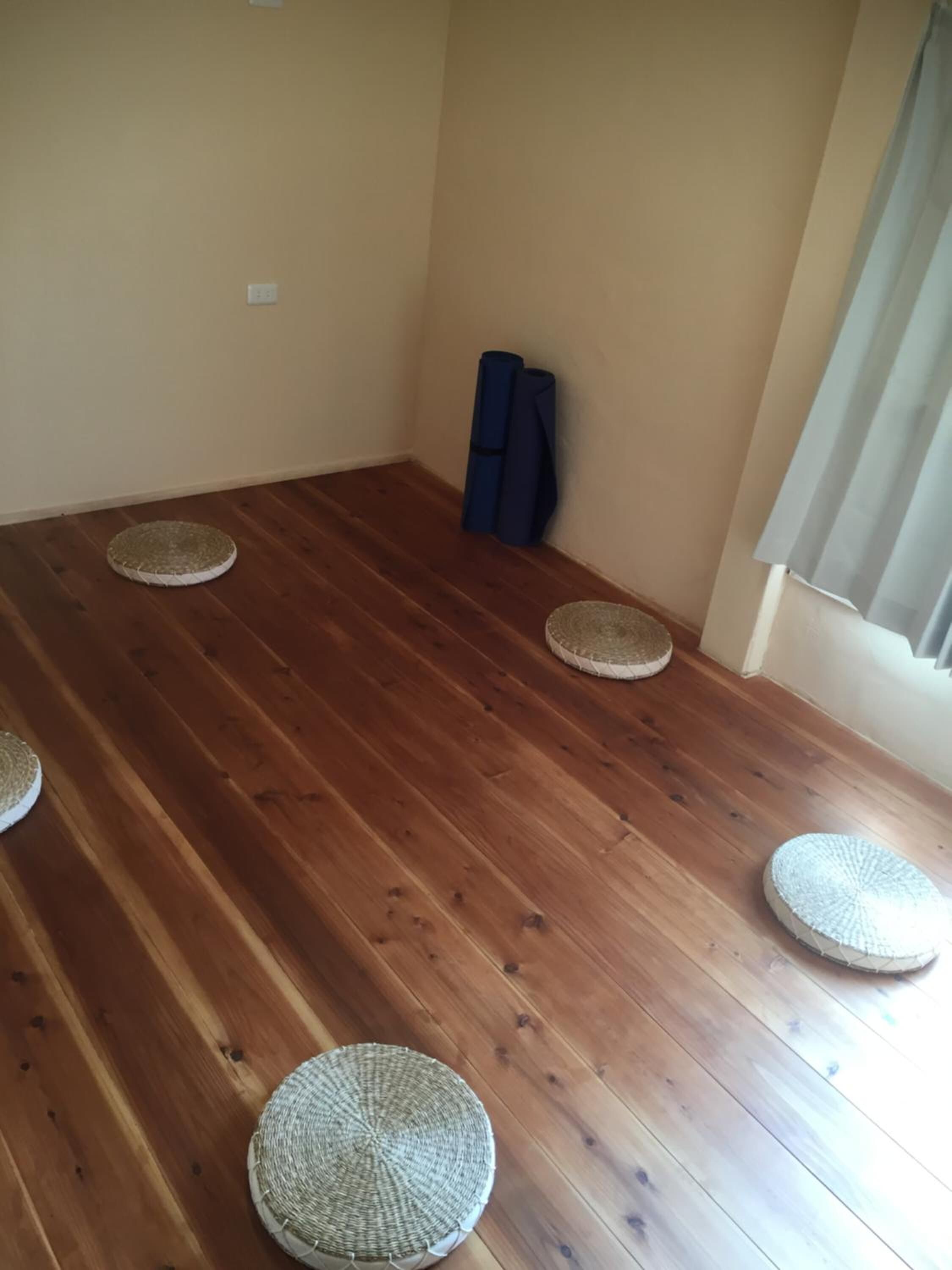 A room with a wooden floor, four woven mats, a blue mat rolled up, and a curtain on the window.