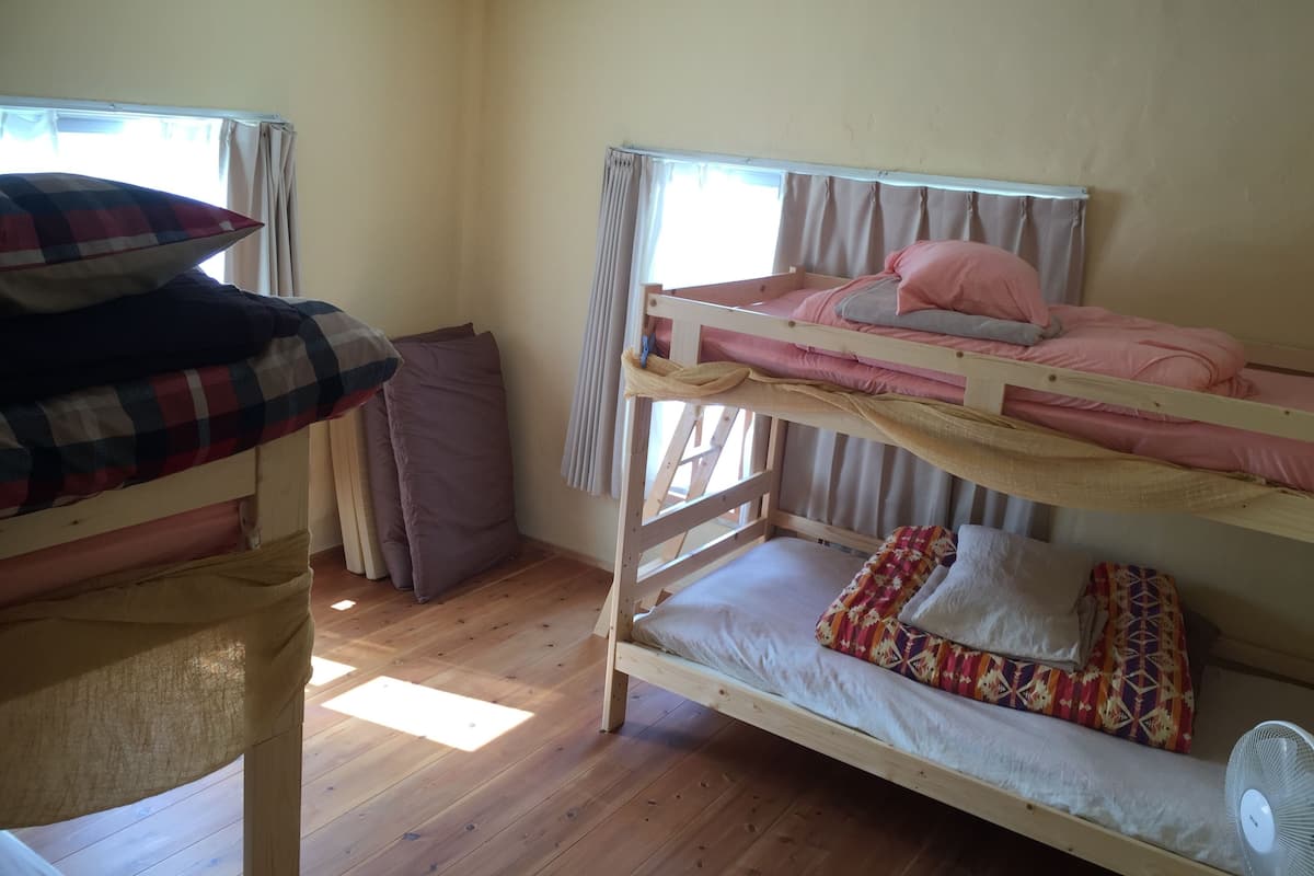 Une chambre avec un lit superposé, un plancher en bois, deux fenêtres avec des rideaux et un ventilateur au-dessus du lit du bas.
