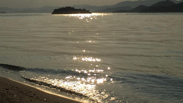 Une plage aux eaux calmes où se reflète la lumière du soleil, une île lointaine et des montagnes en arrière-plan.