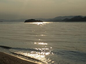 Ein Strand mit ruhigem Wasser, das Sonnenlicht reflektiert, eine ferne Insel und Berge im Hintergrund.