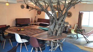 Un espace repas avec une grande table en bois entourée de chaises, une sculpture rustique en forme d’arbre servant de centre de table, et un canapé en arrière-plan.