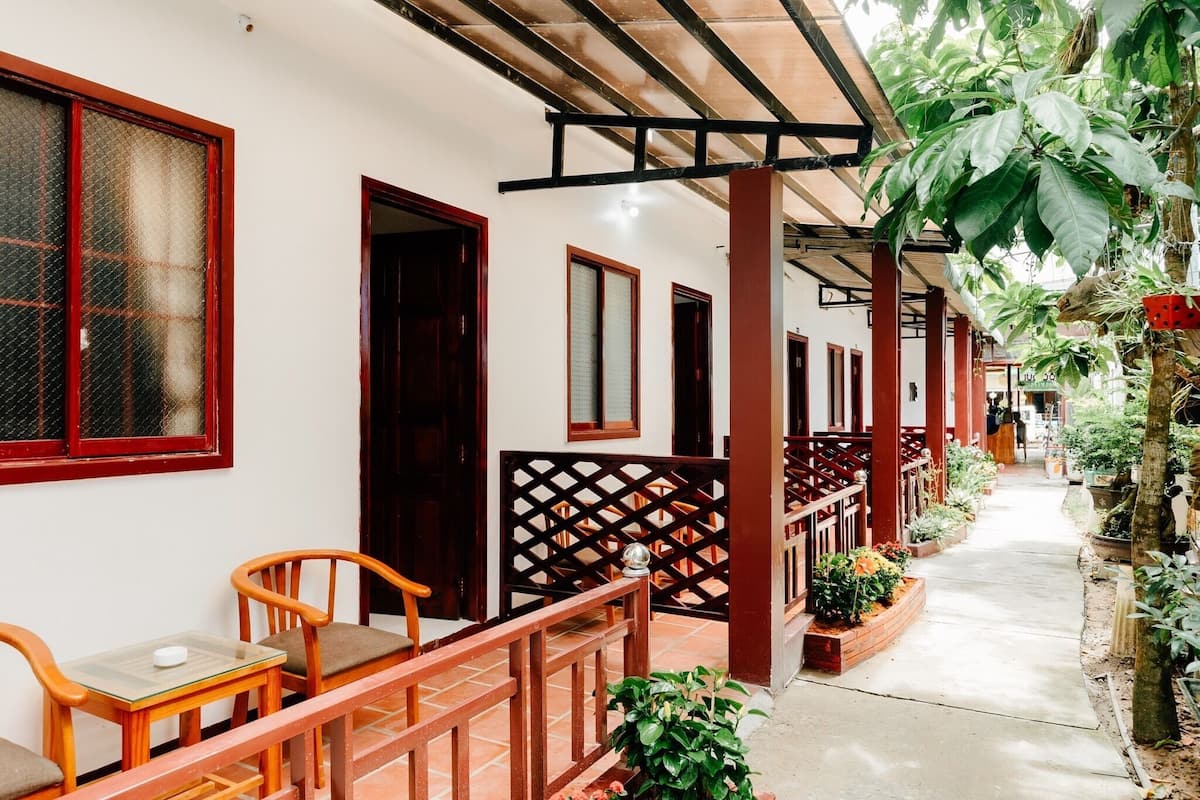 A covered outdoor area with wooden chairs and tables, a pathway lined with potted plants, and a building with multiple doors and windows.