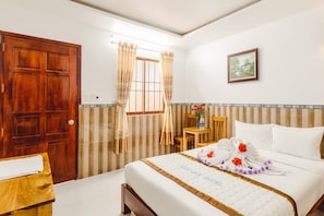 A hotel room with a wooden door, a bed with a white bedspread and floral decorations, a small wooden table with a vase, and a window with curtains. - Ngoc Qui Bungalow (Phu Quoc)