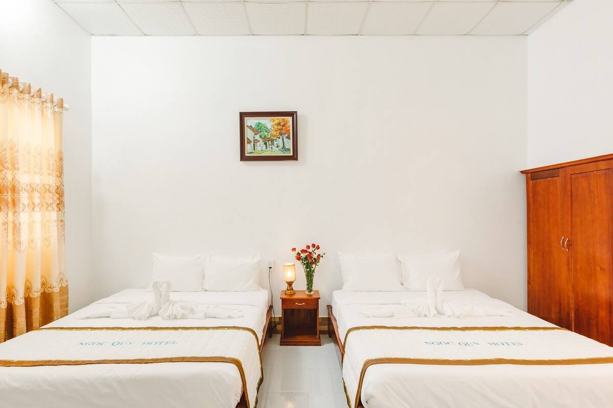 A hotel room with two beds, a nightstand with a lamp and flowers, and a framed picture on the wall.