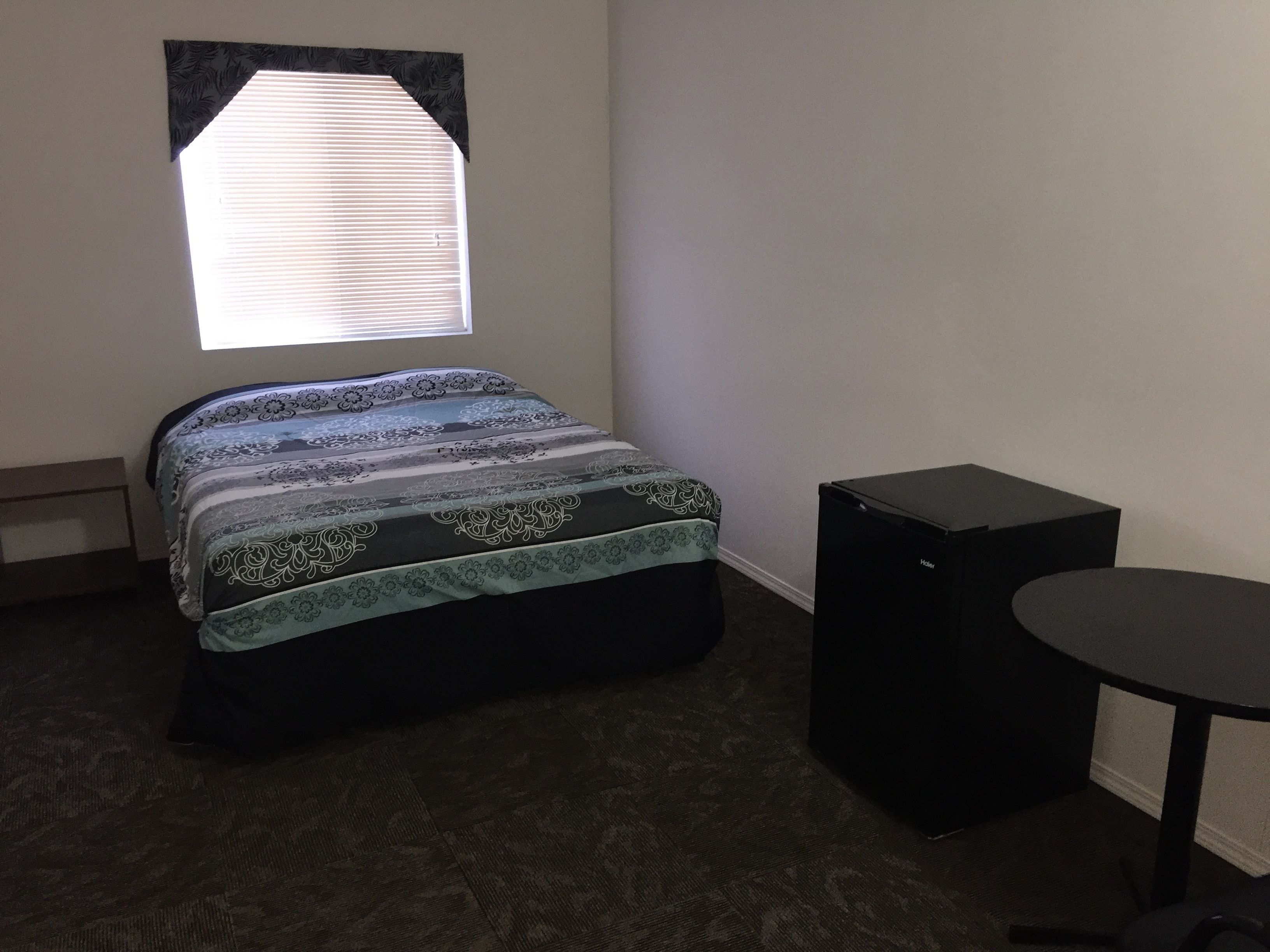Un dormitorio con cama, una ventana con persianas, un altavoz negro pequeño y una mesa redonda.