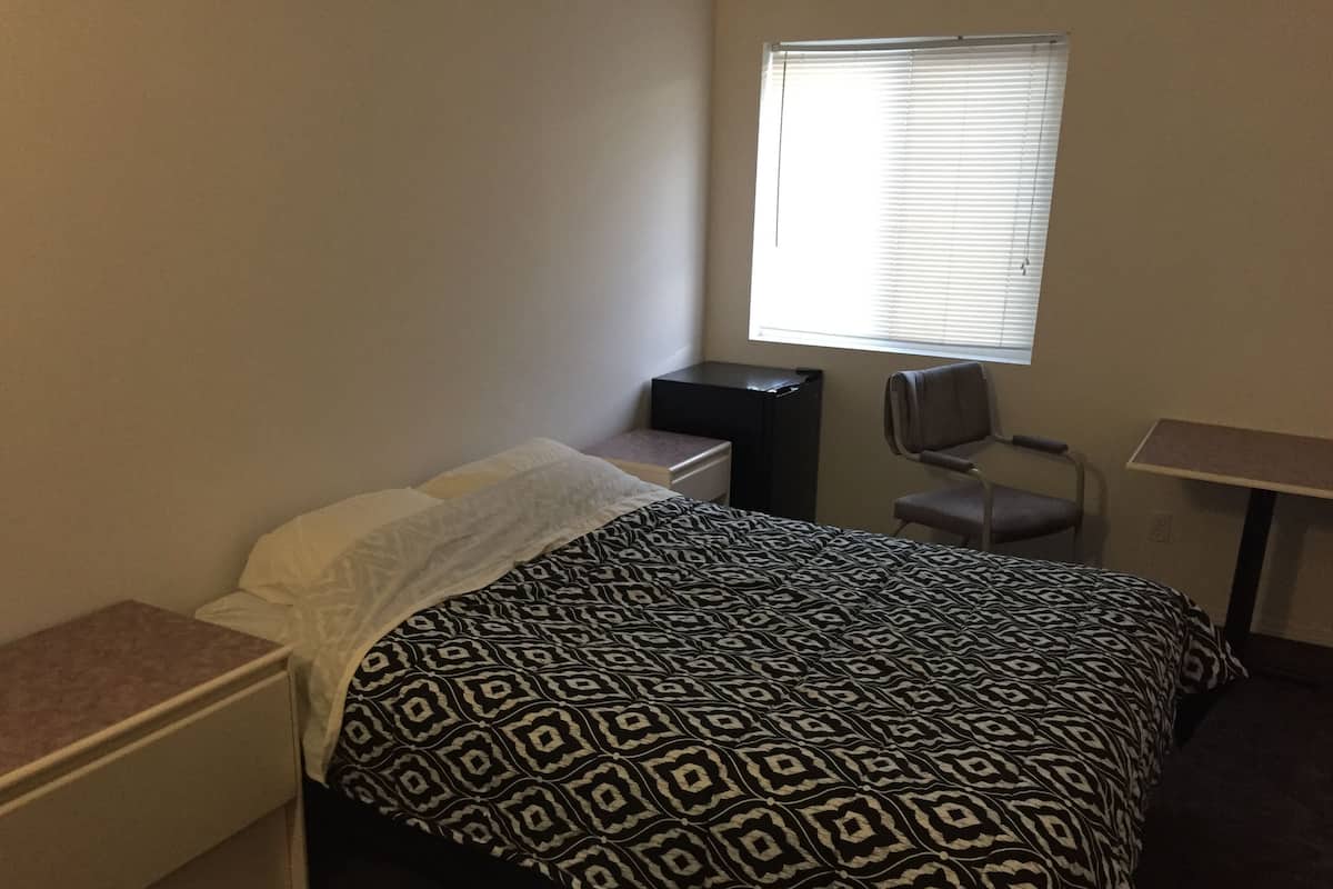 Um quarto pequeno e modesto, com uma cama, uma escrivaninha, uma cadeira e uma janela.