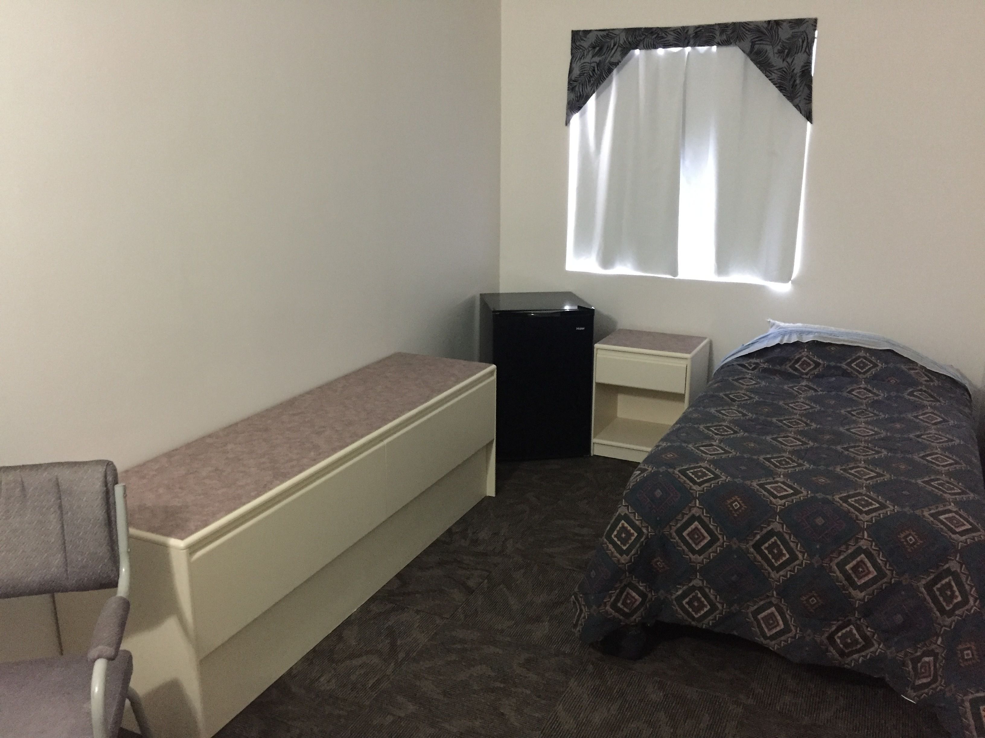 Une petite pièce simple, avec un lit, un bureau, une chaise, une petite table et une fenêtre avec un rideau.