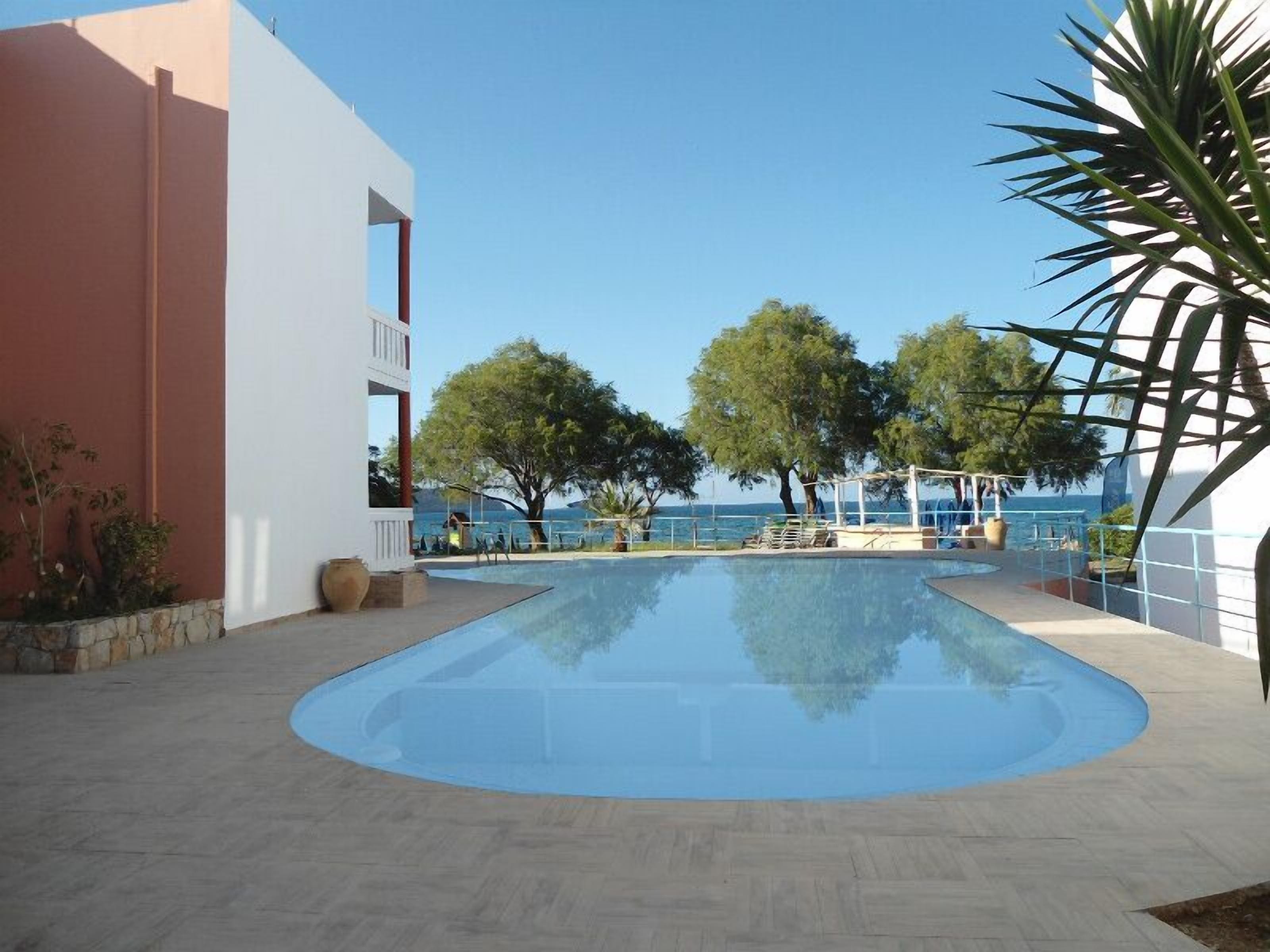 A swimming pool with a clear view of the sea, surrounded by trees and a building with a balcony.