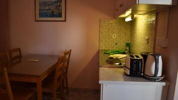 Una cocina pequeña con una mesa, sillas, un tostador, una tetera y un escurreplatos verde.