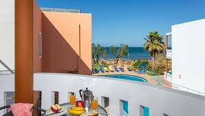 Un balcón con una mesa lista para el desayuno, con vista a una piscina y una playa con sillones de descanso y palmeras.