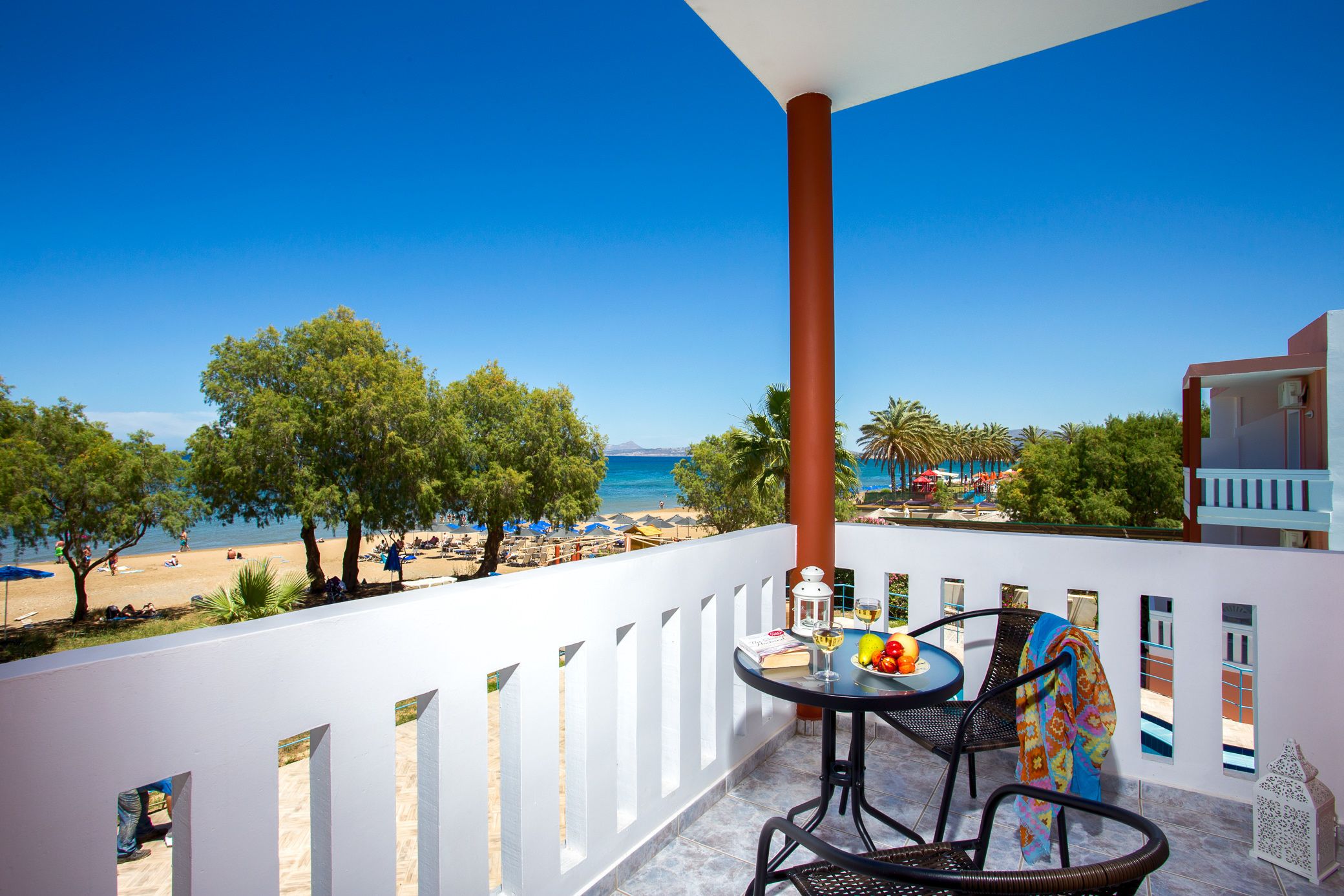 A balcony with a table and chairs, overlooking a beach with palm trees and a clear blue sky.