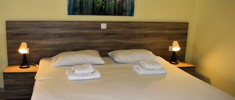 Una cama bien tendida con sábanas blancas y dos almohadas, con dos mesitas de noche y lámparas a ambos lados, en una habitación con cabecera de madera y un cuadro abstracto y colorido en la pared.