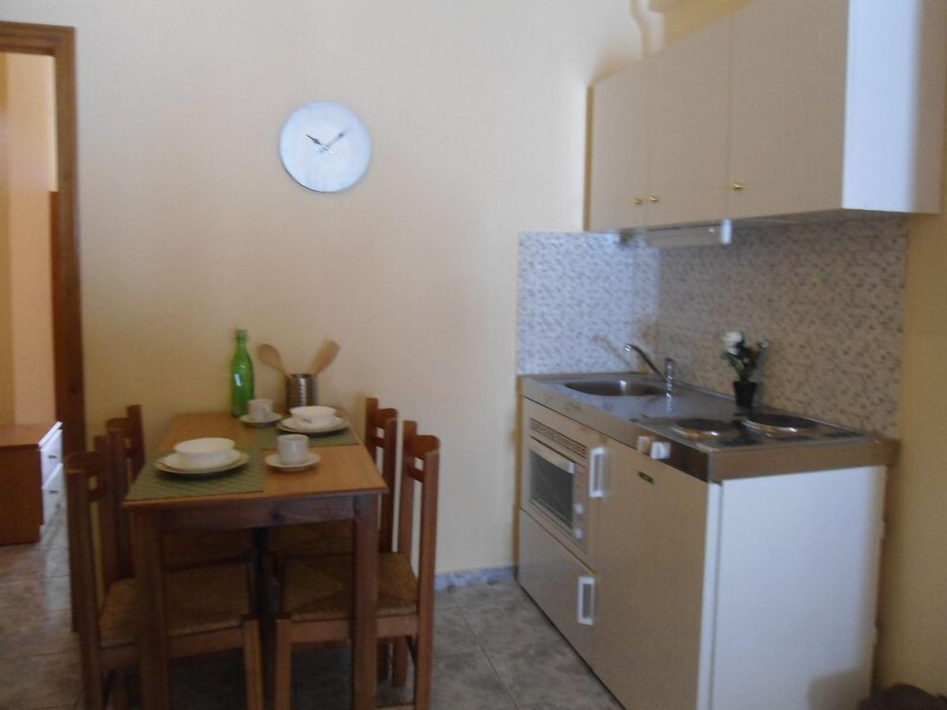 Una cocina pequeña con una mesa de madera, sillas, un reloj y electrodomésticos básicos.