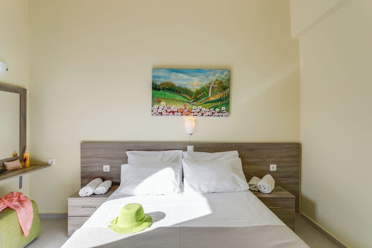 Une chambre d’hôtel avec un lit, une table de chevet, un fauteuil vert, un tableau au mur et une lampe.