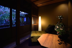 Una stanza tradizionale giapponese con pavimento in tatami, un tavolino in legno, un’area salotto con cuscini e vista su un giardino attraverso un paravento shoji.