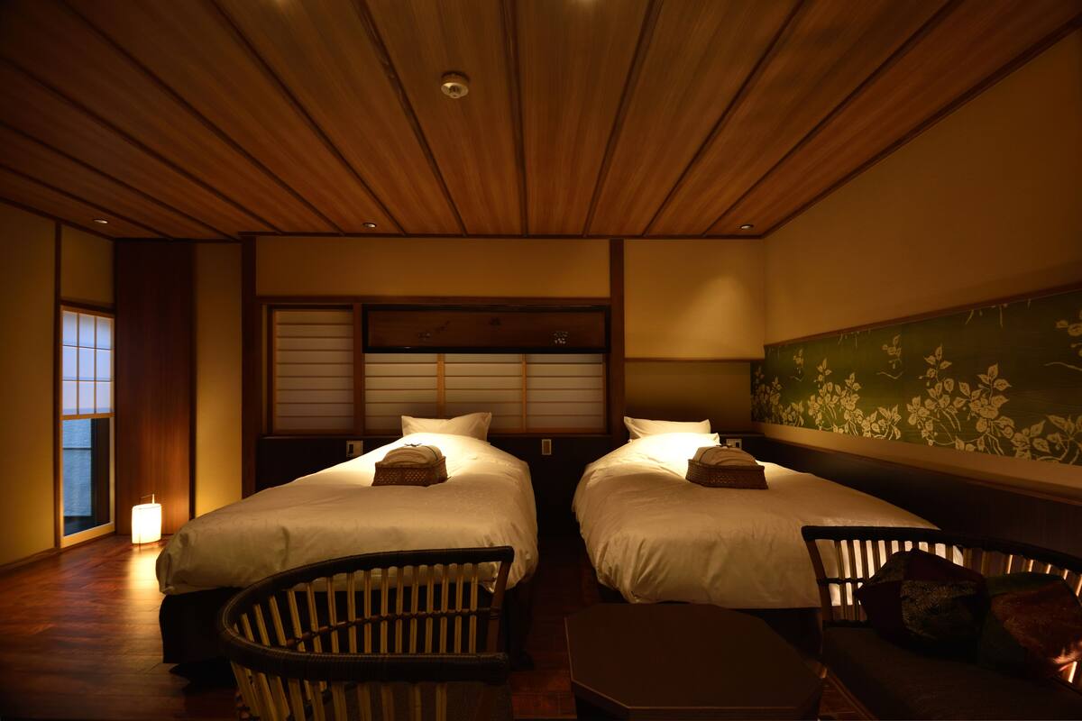 A hotel room with two beds, a wooden ceiling, a green floral wall, and a traditional Japanese sliding door.