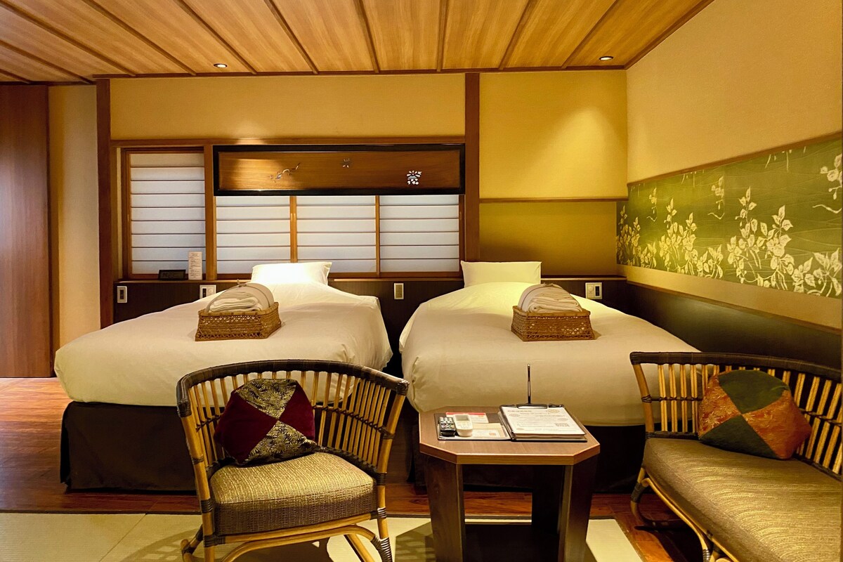 Habitación de hotel con dos camas, una zona de estar con una silla y una mesa, y una pared de estilo japonés tradicional.