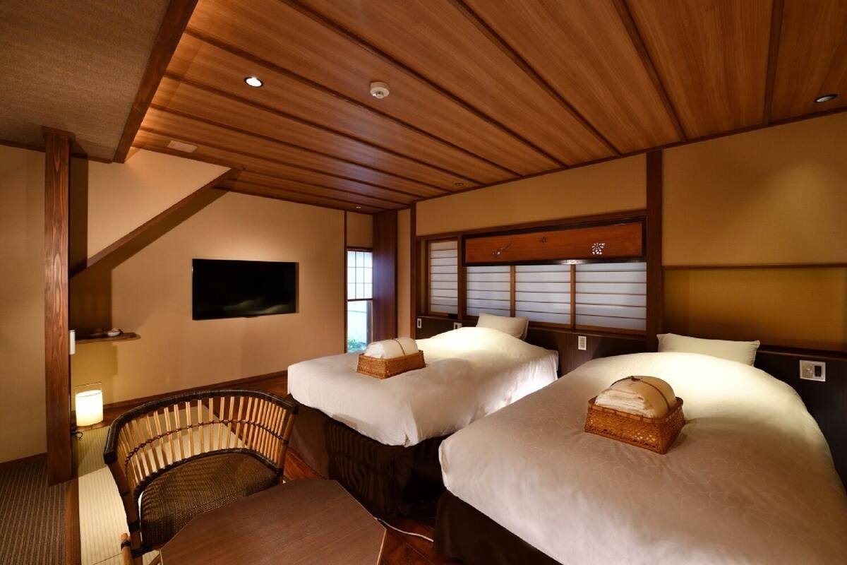 A hotel room with two beds, a wooden ceiling, a television, and a traditional Japanese-style chair.