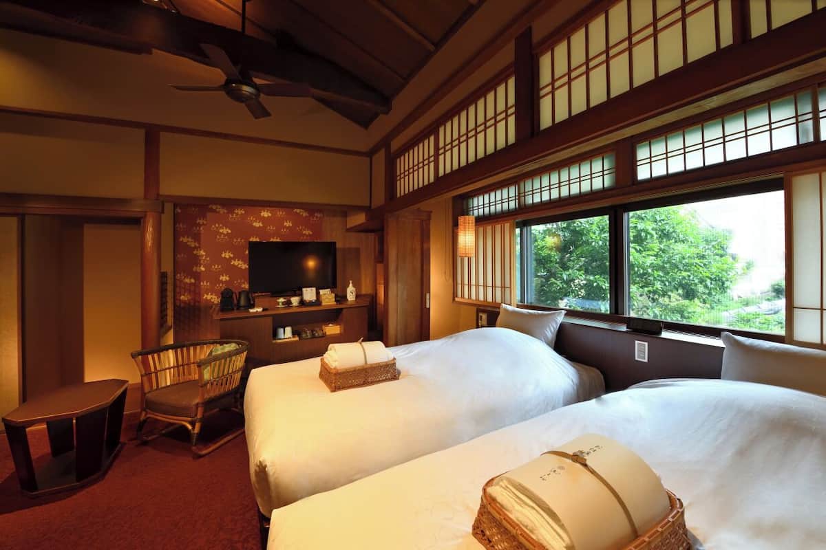 A traditional Japanese-style room with two beds, a television, a wooden chair, and a window with a view of greenery.