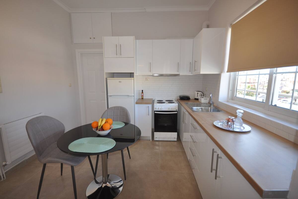 A compact kitchen with white cabinets, a dining table with chairs, a built-in oven, and a window with a view.
