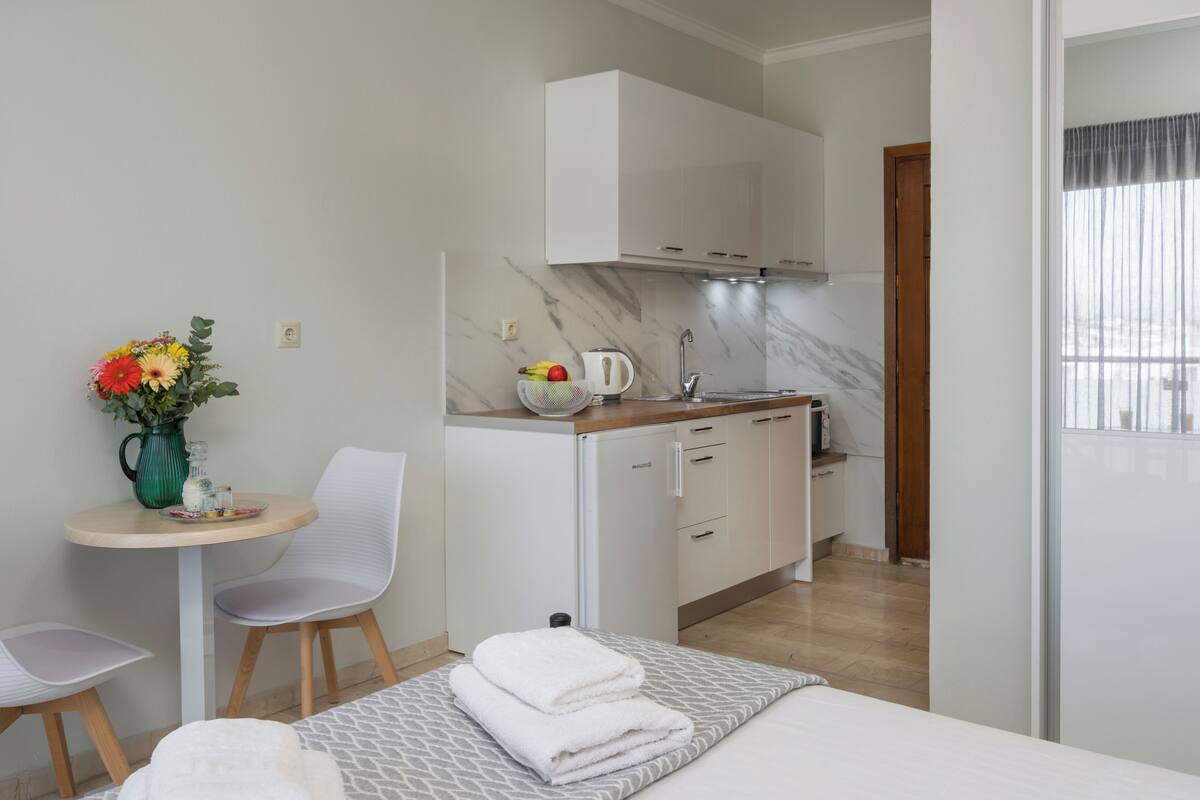 A compact hotel room with a kitchenette, a dining area with a table and chairs, a bed with a gray patterned bedspread, and a vase with flowers on the side table.