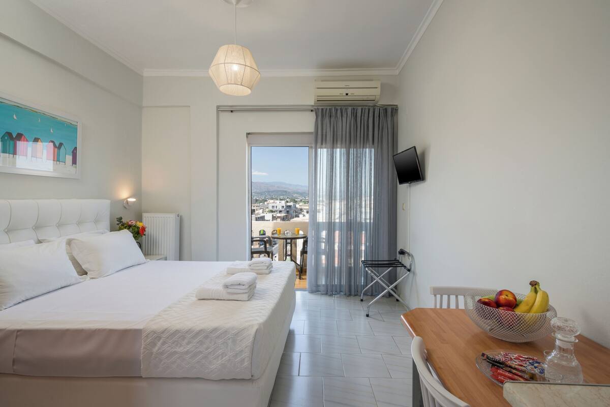 A hotel room with a bed, a dining table with a fruit bowl, a TV mounted on the wall, and a view through the window.