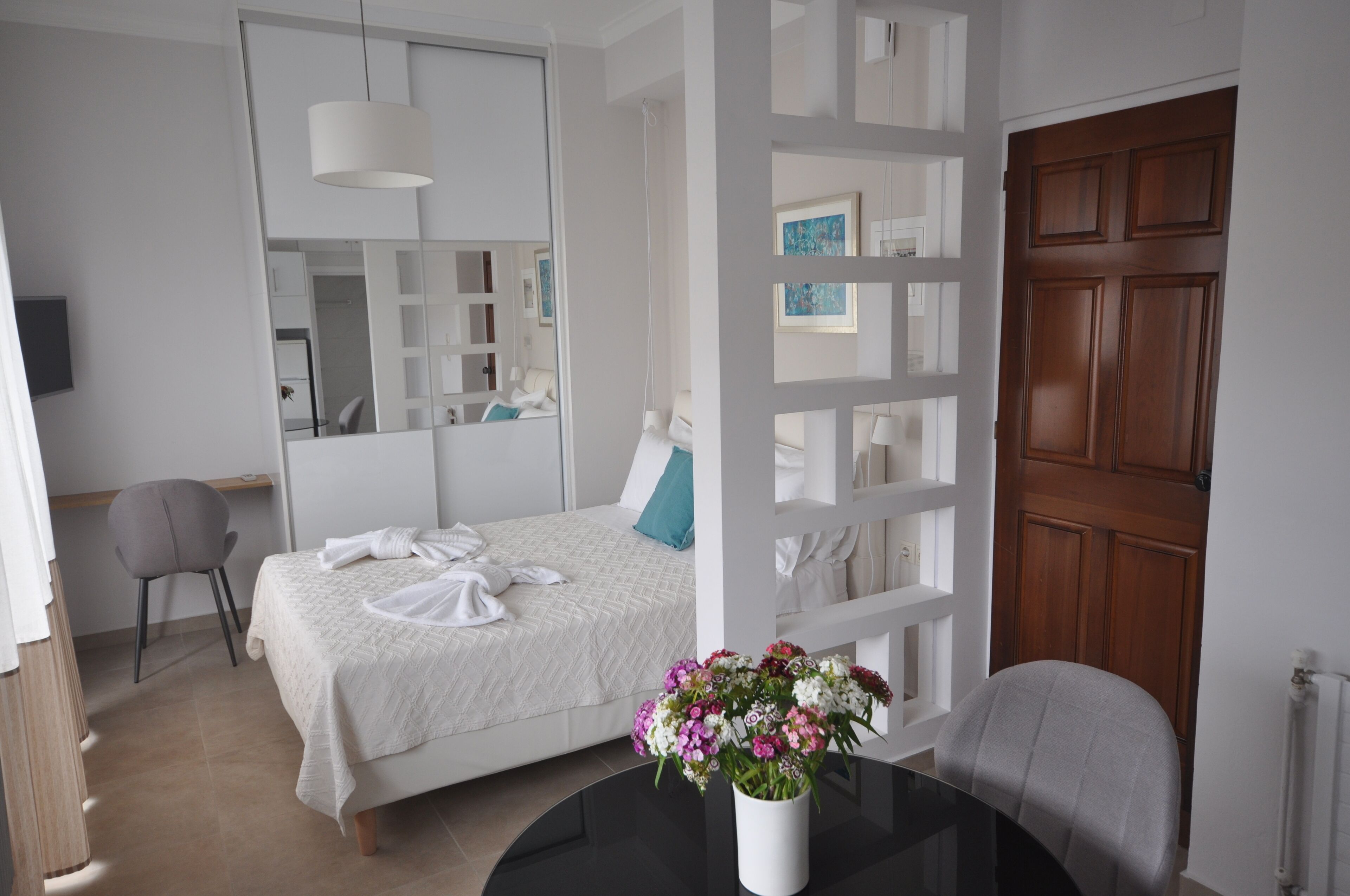 A hotel room with a bed, a chair, a table with a vase of flowers, a wooden door, and a white wardrobe.