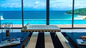 Une pièce dotée d’une grande fenêtre donnant sur une plage et l’océan, d’un coin salon confortable et d’une table sur laquelle sont posés des livres et un chandelier.