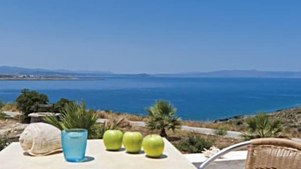 Una mesa con una bebida, una manzana y una concha marina, con vista a la costa y montañas al fondo.