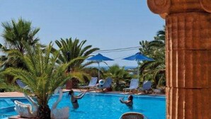 Zona de piscina con sillones de descanso y sombrillas, rodeada de palmeras.