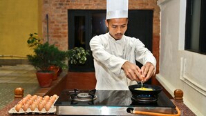 Un chef prepara comida en una cocina con pared de ladrillo y plantas en macetas.