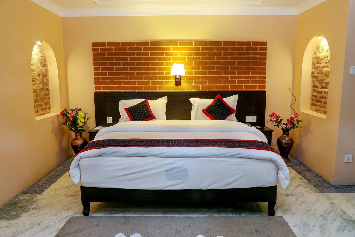 A neatly arranged hotel room with a brick-patterned headboard, a large bed with white bedding and red accents, two bedside tables with lamps, and a vase with flowers on each side.
