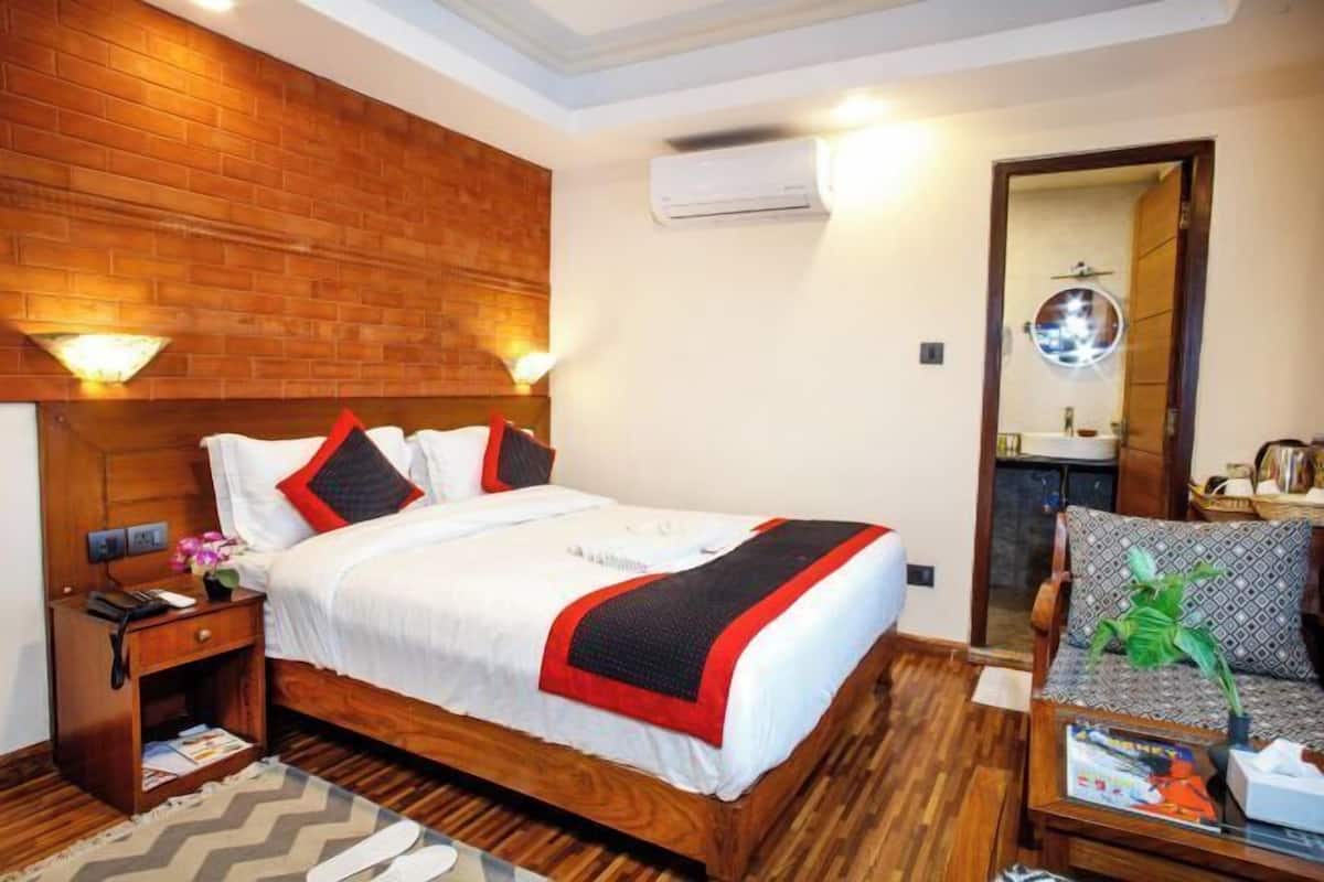 A hotel room with a wooden headboard, a bed with white bedding and red accents, a nightstand with a lamp, and a wooden floor.