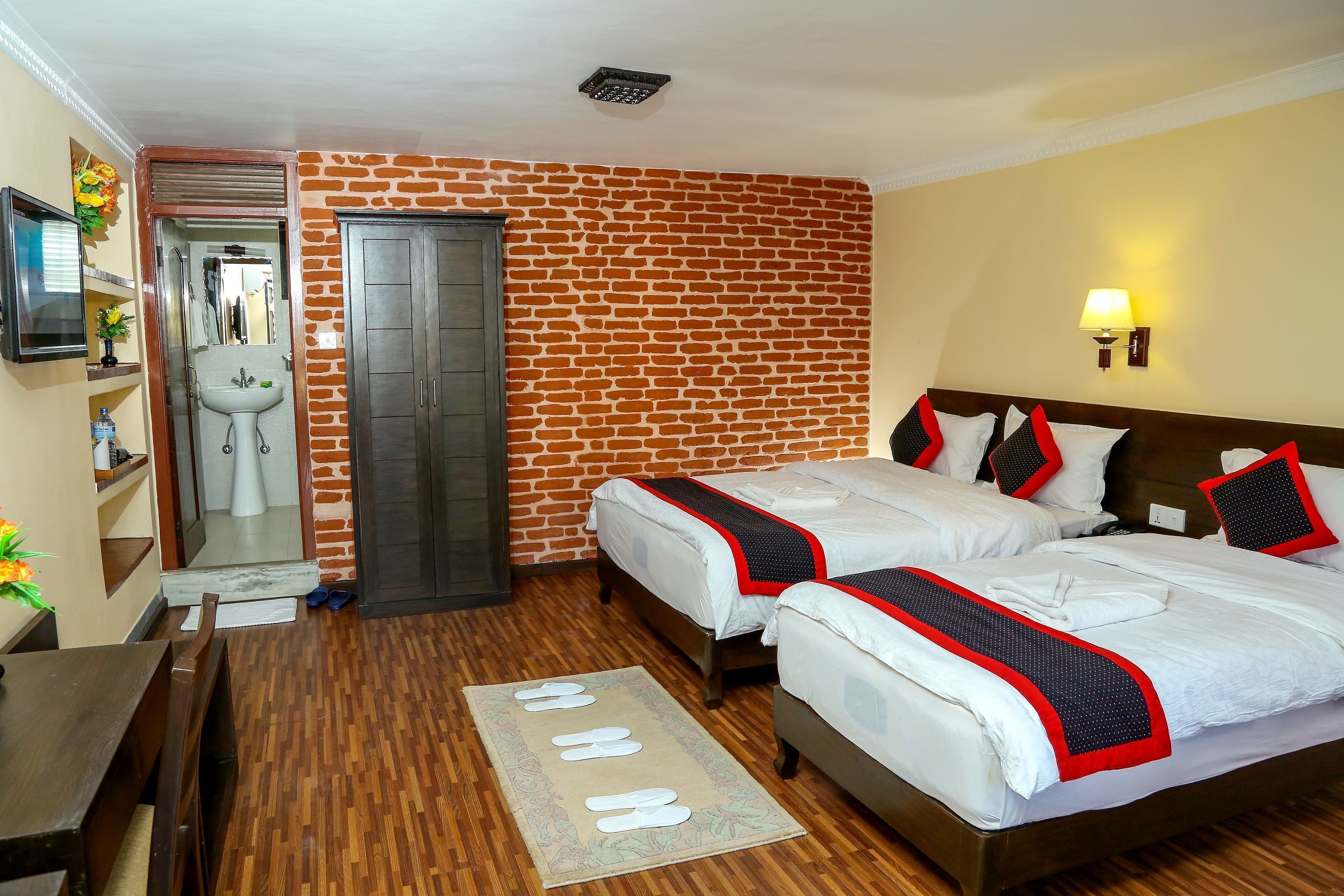 Habitación de hotel con dos camas, piso de madera, pared con diseño de ladrillo, un lavamanos y un televisor.