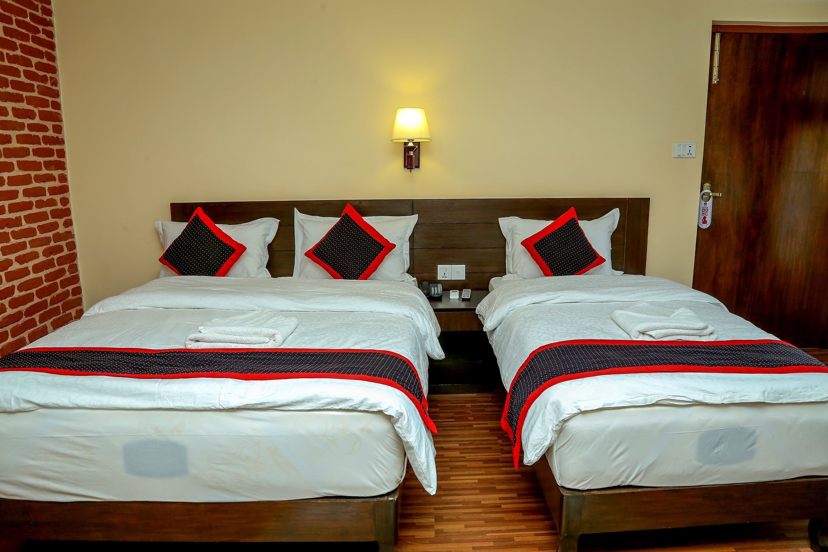 A hotel room with two beds, each with white bedding and red and black pillows. There is a wall-mounted lamp above the beds, and a wooden door on the right.