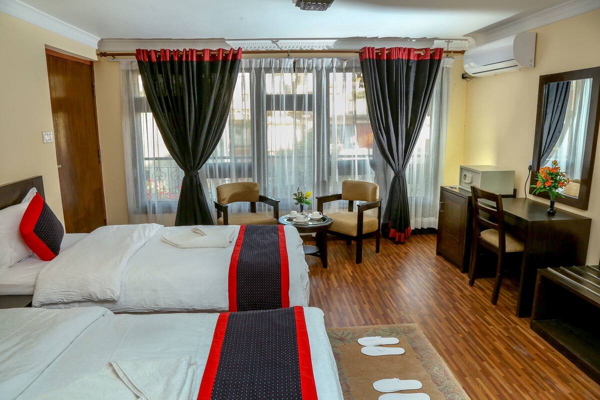 A hotel room with two beds, a desk, a chair, a small table with a plant, and an air conditioning unit.