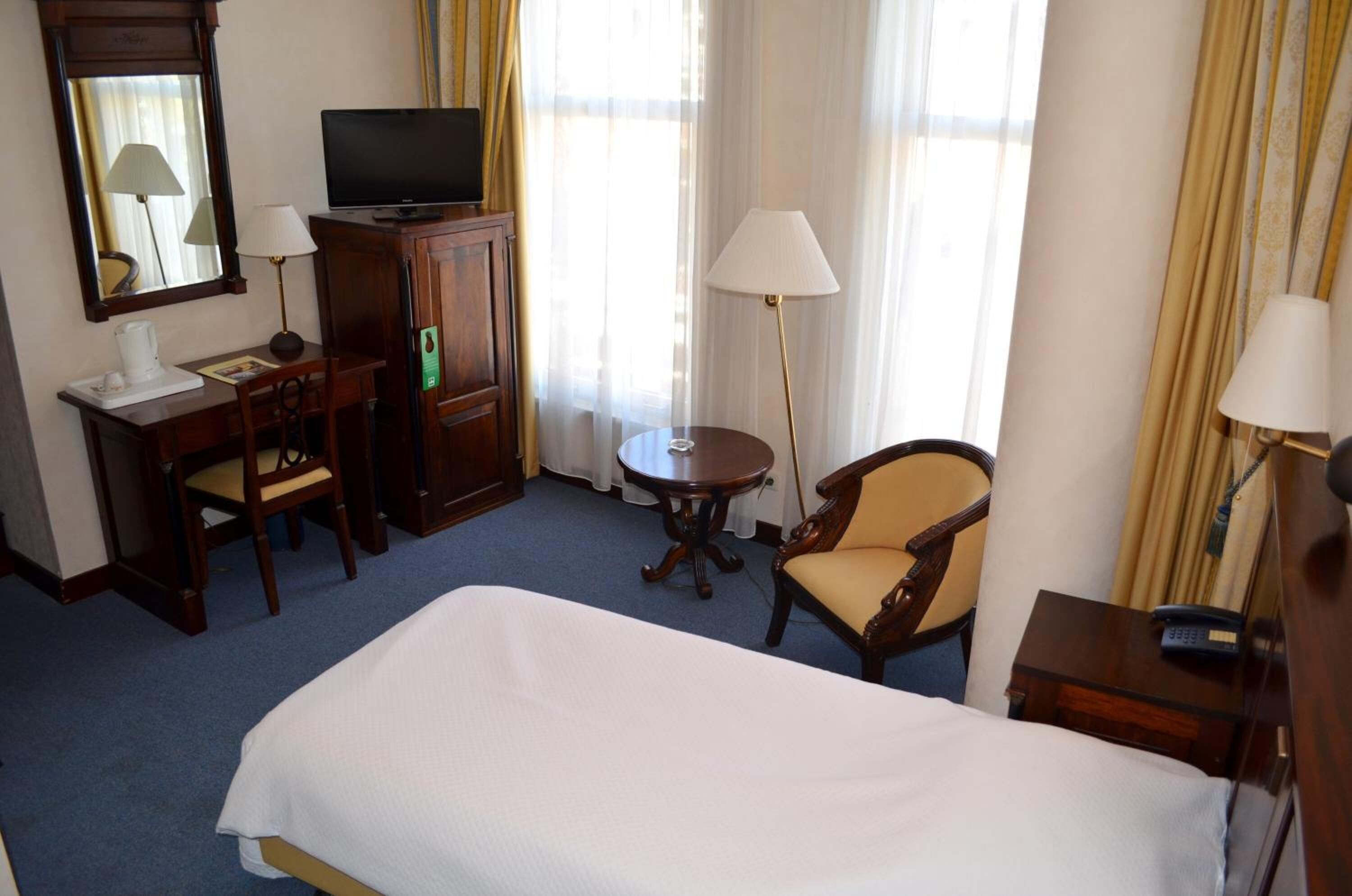 A hotel room with a bed, a desk with a television, a chair, a small table, and a lamp.