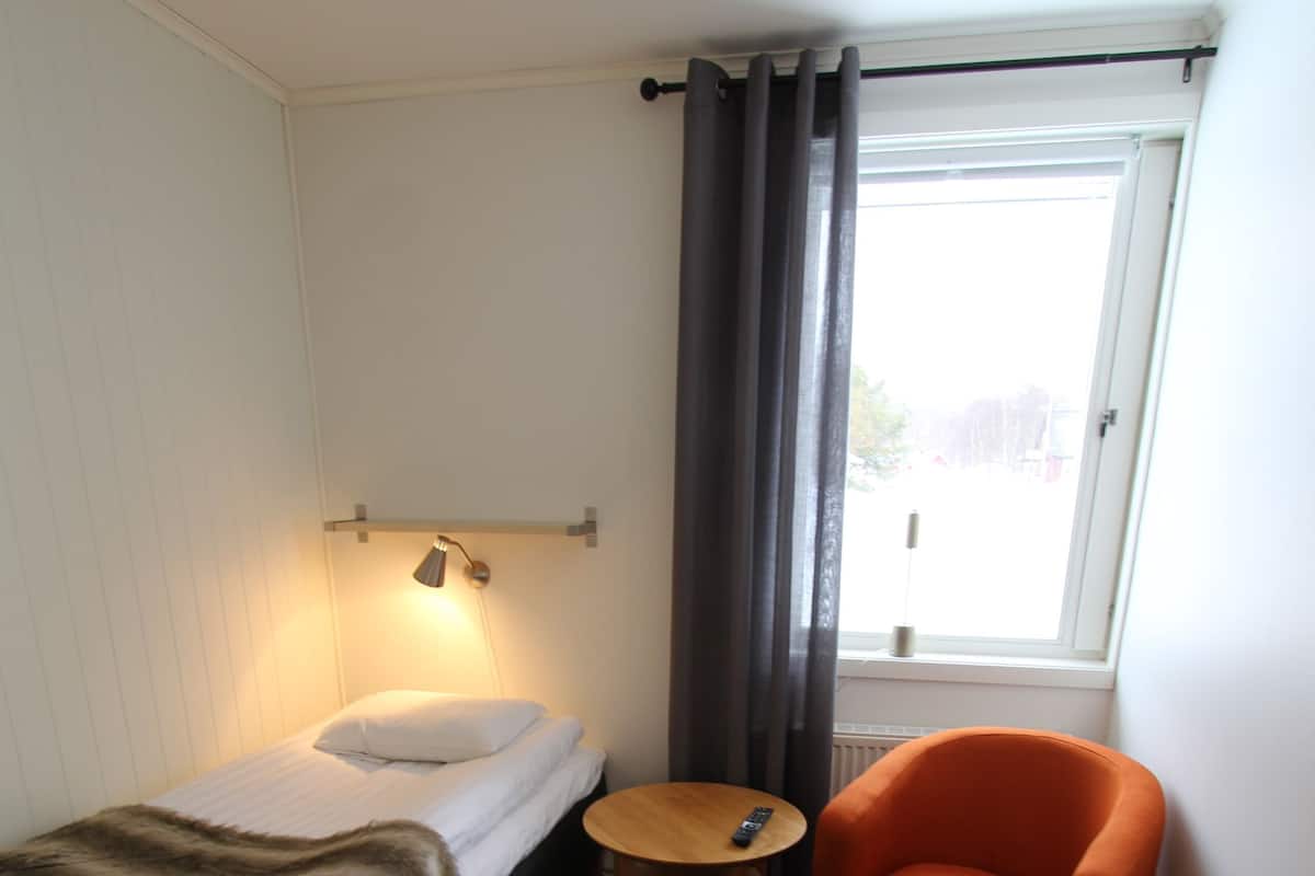 Una habitación de hotel pequeña con una cama, una silla, una mesita, una lámpara, una ventana con cortinas y un radiador.