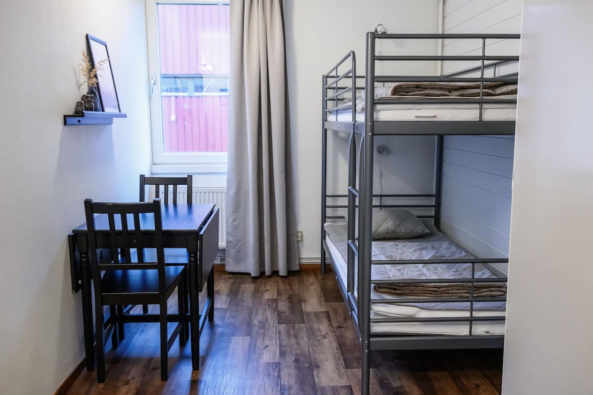 Habitación con literas, suelo de madera, una mesa con sillas y una ventana con cortinas.