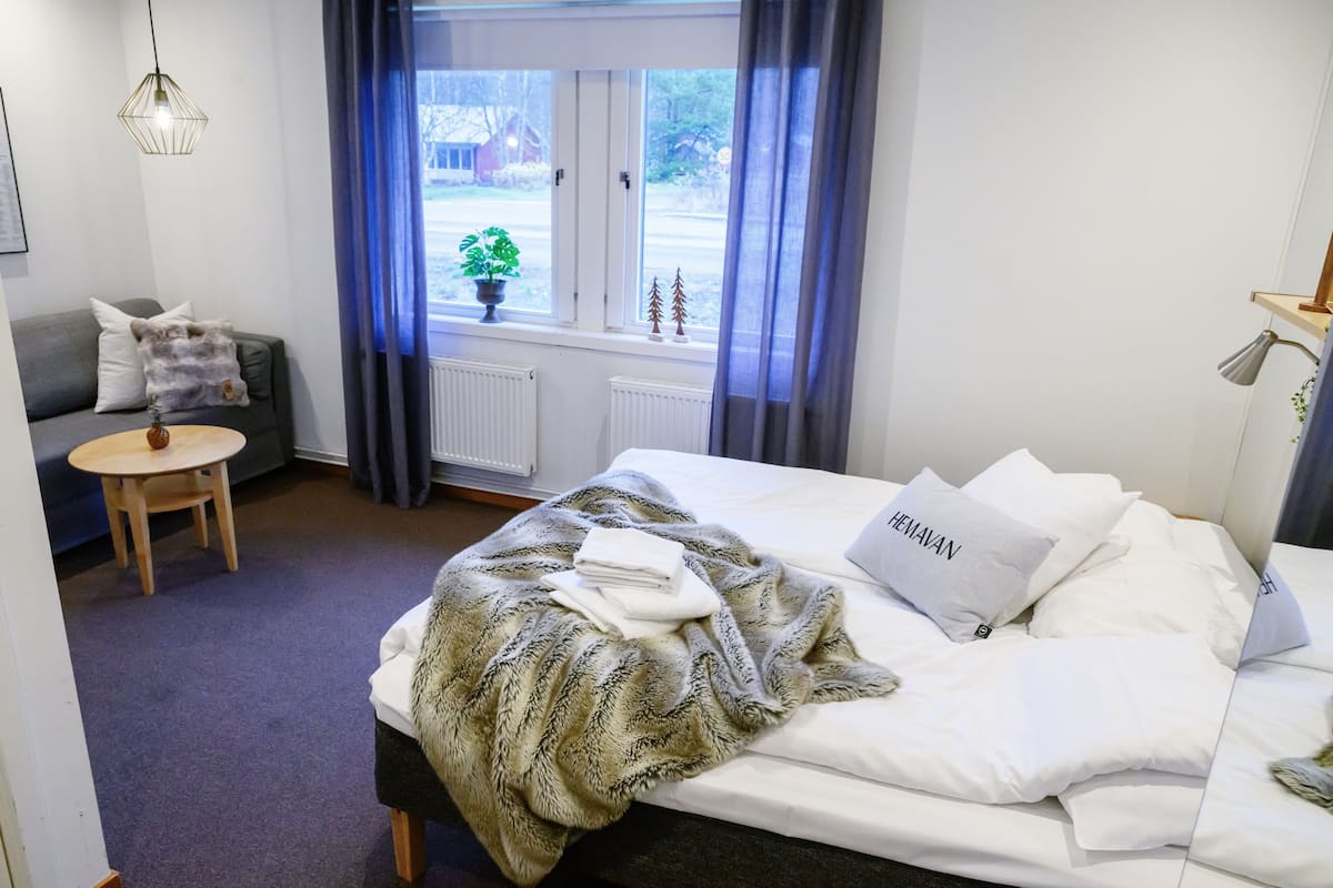 Una habitación de hotel con cama, una mesita pequeña con una silla, un sofá, una ventana con cortinas y una planta en el alféizar.
