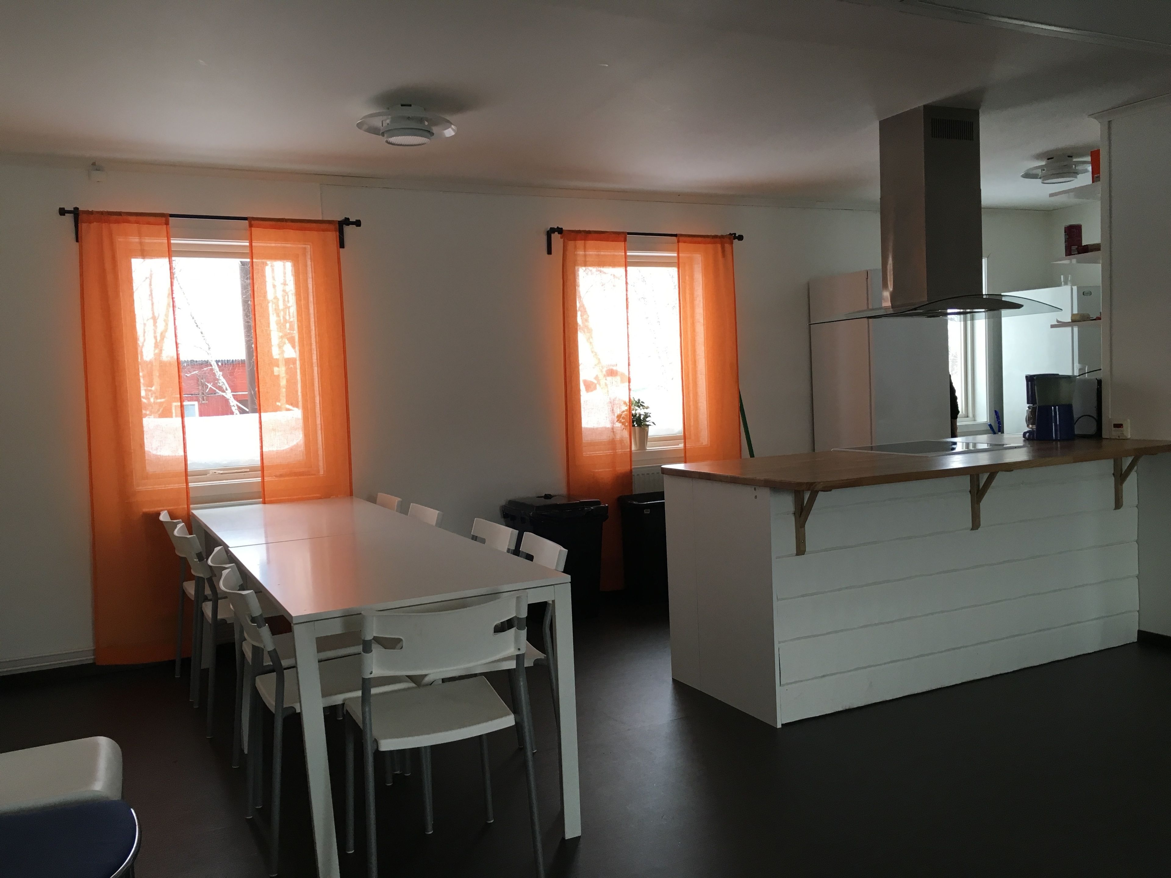 Una cocina compacta con una isla central, muebles blancos y un ventanal con cortinas naranjas.