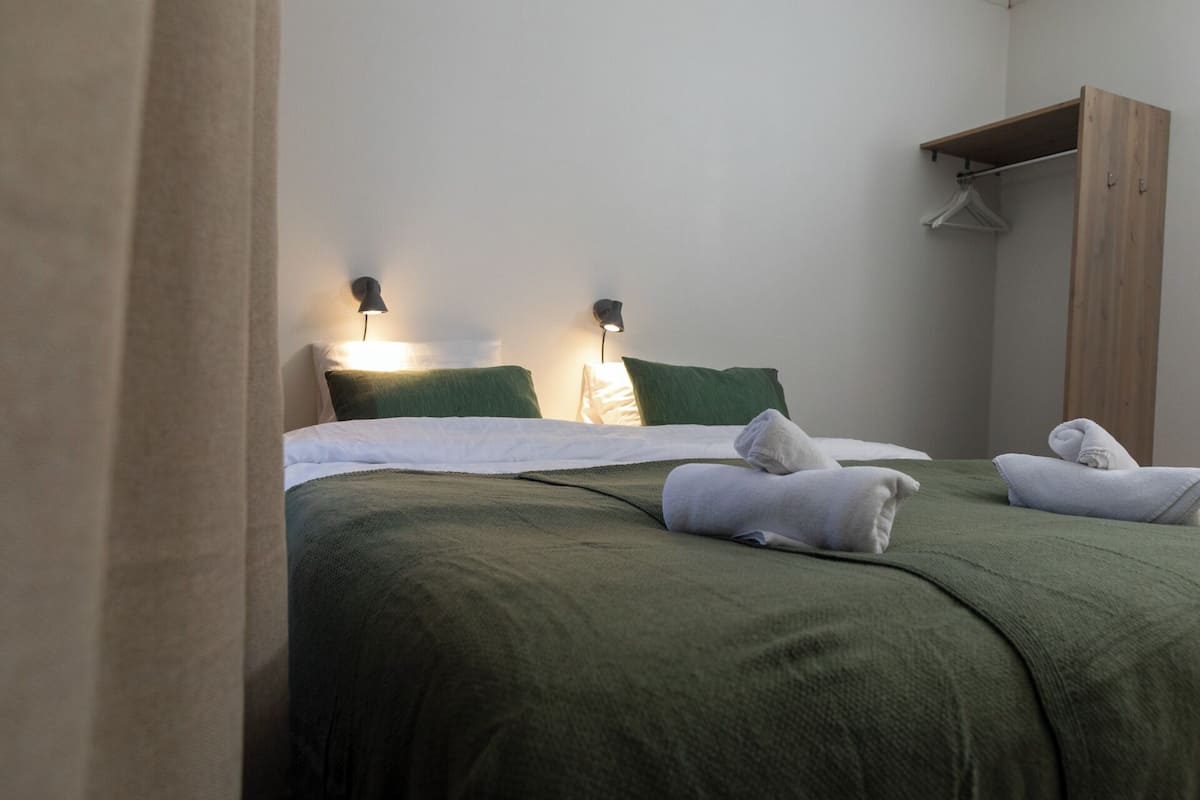 Una cama bien hecha con ropa de cama verde, dos lámparas de noche y un armario de madera con perchero.
