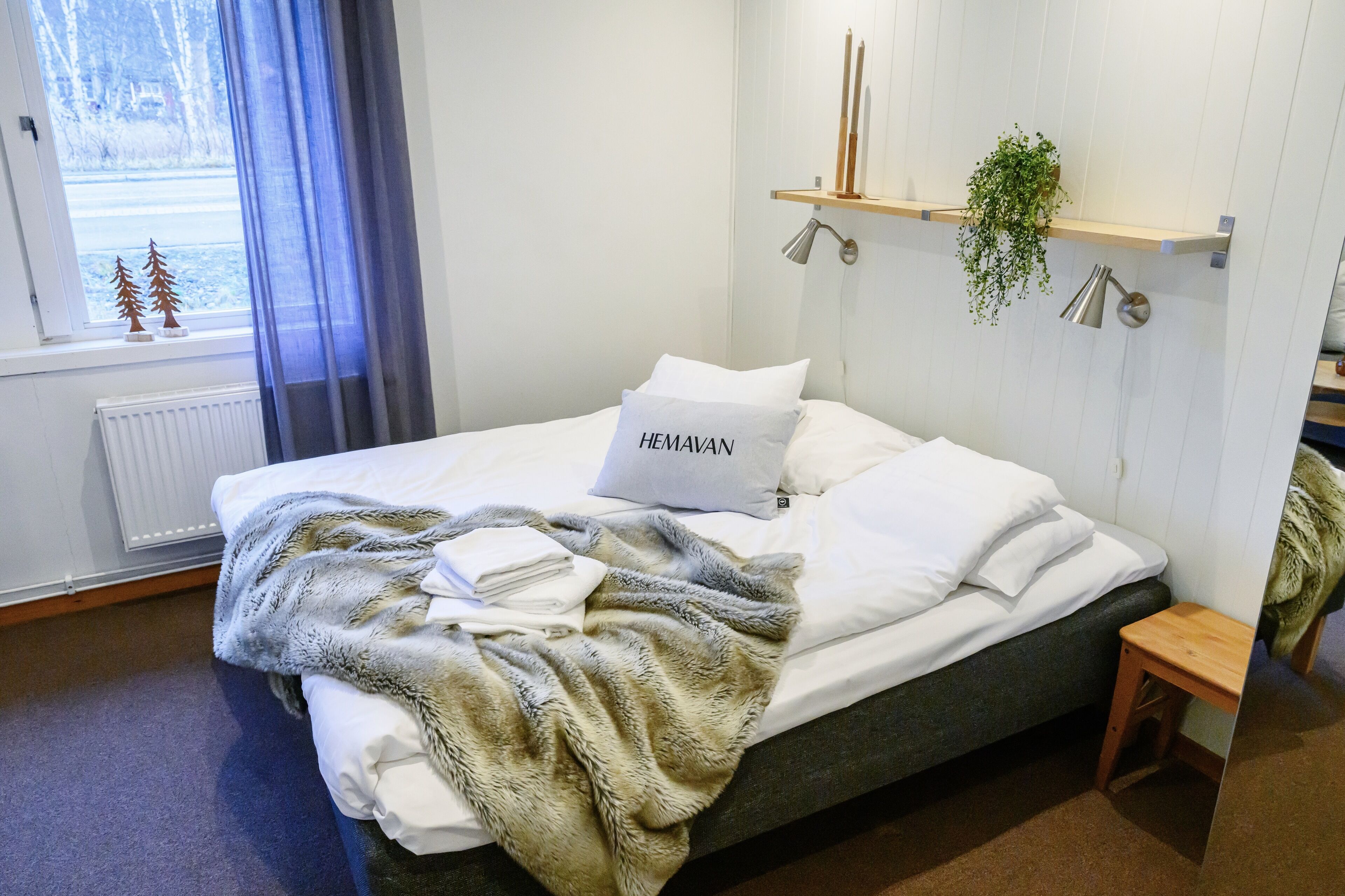Een net opgemaakt bed met een zachte deken en een kussen met de tekst 'HEMAVAN' in een moderne kamer met een raam, radiator en een plank met een plantje.