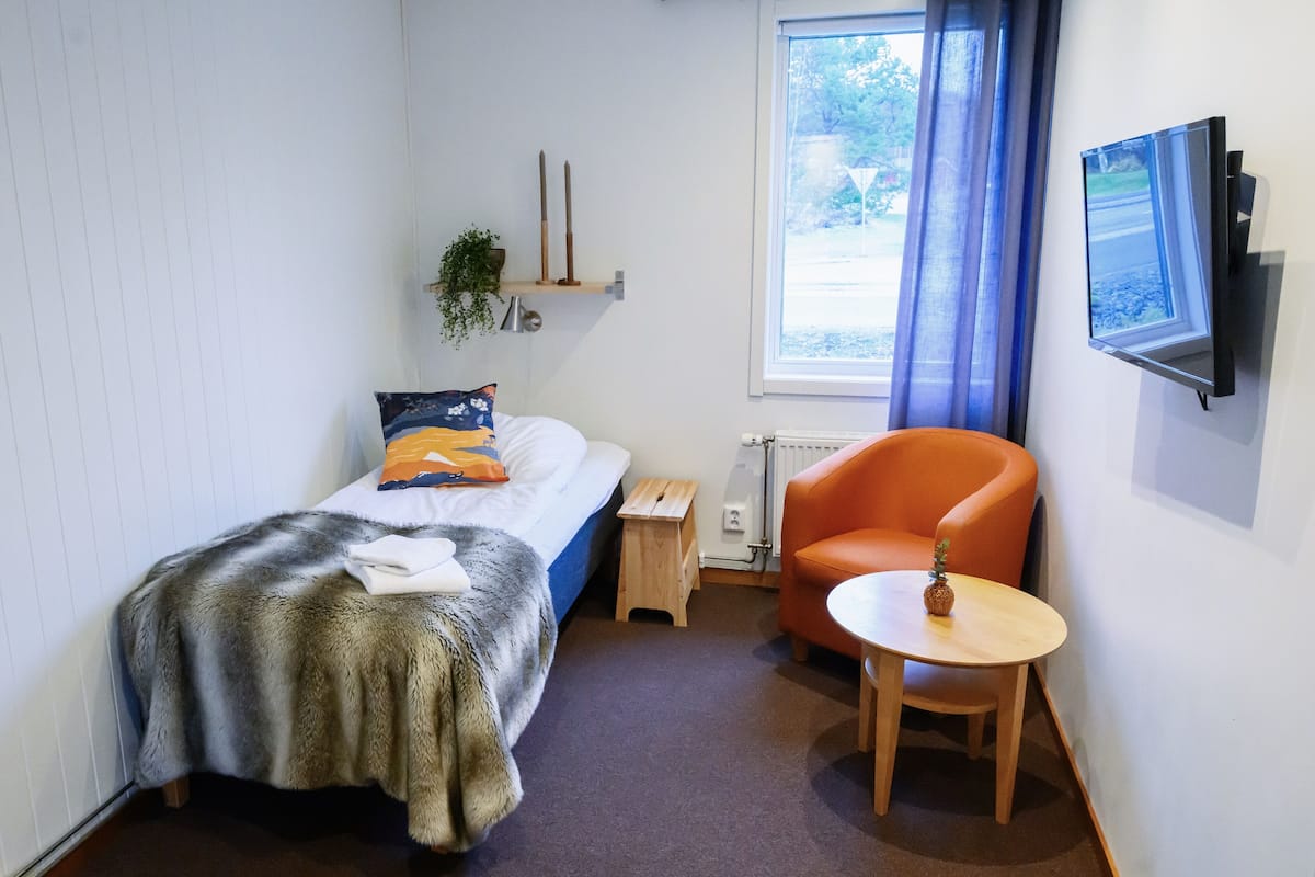 Una habitación pequeña y ordenada con una cama, una silla, una mesita, un televisor y una ventana con cortinas.