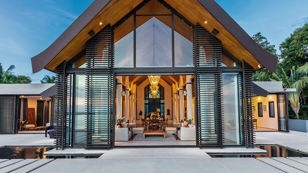 Une maison moderne dotée d’une grande façade en verre, d’un toit en bois et d’un espace de salle à manger central agrémenté d’un lustre.