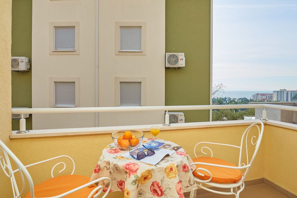 A balcony with a table set for two, featuring a floral tablecloth, a bowl of oranges, and a glass of orange juice.