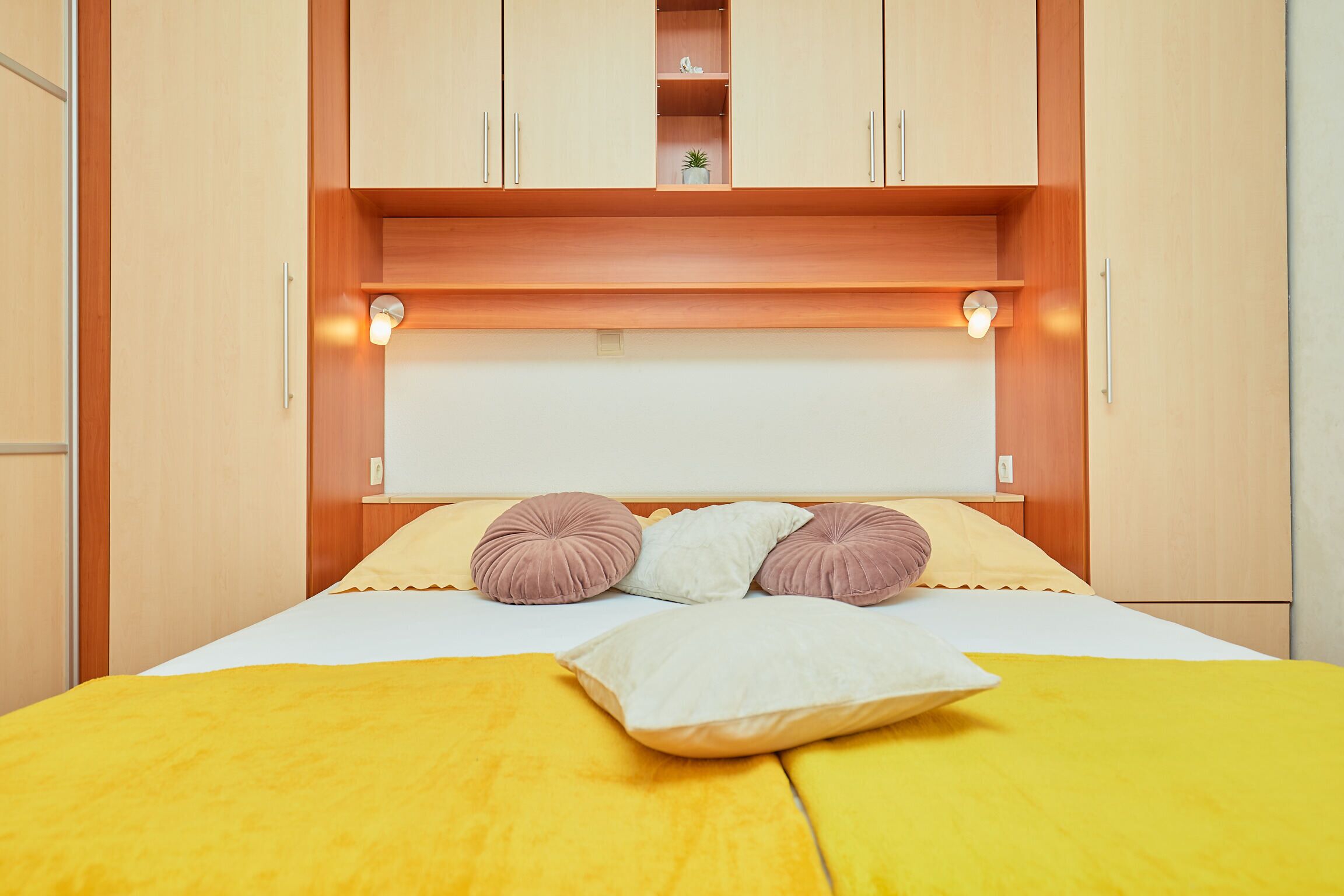 Een net opgemaakt bed met een gele deken en meerdere kussens, een houten hoofdbord met ingebouwde verlichting, en een kleine plant op een plank boven het bed.