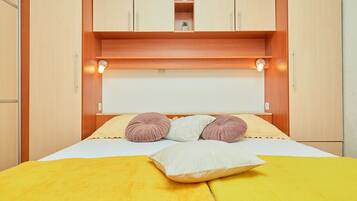 Een net opgemaakt bed met een gele deken en meerdere kussens, een houten hoofdbord met ingebouwde verlichting, en een kleine plant op een plank boven het bed.
