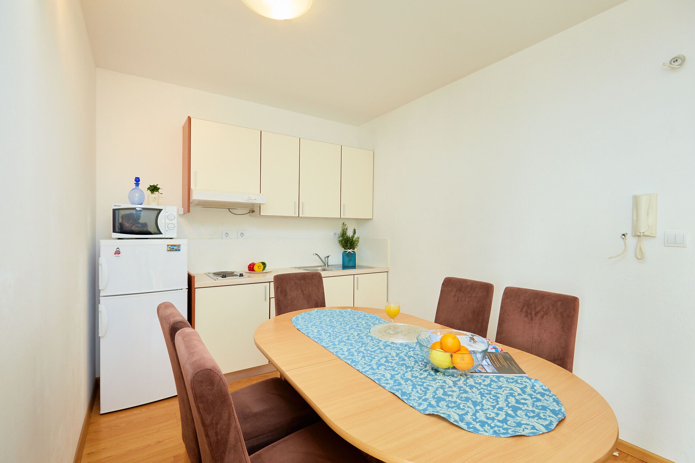 Cocina compacta con microondas, refrigerador y lavamanos. Una mesa de comedor con sillas dispuesta para cuatro personas, y un florero con flores sobre la encimera.