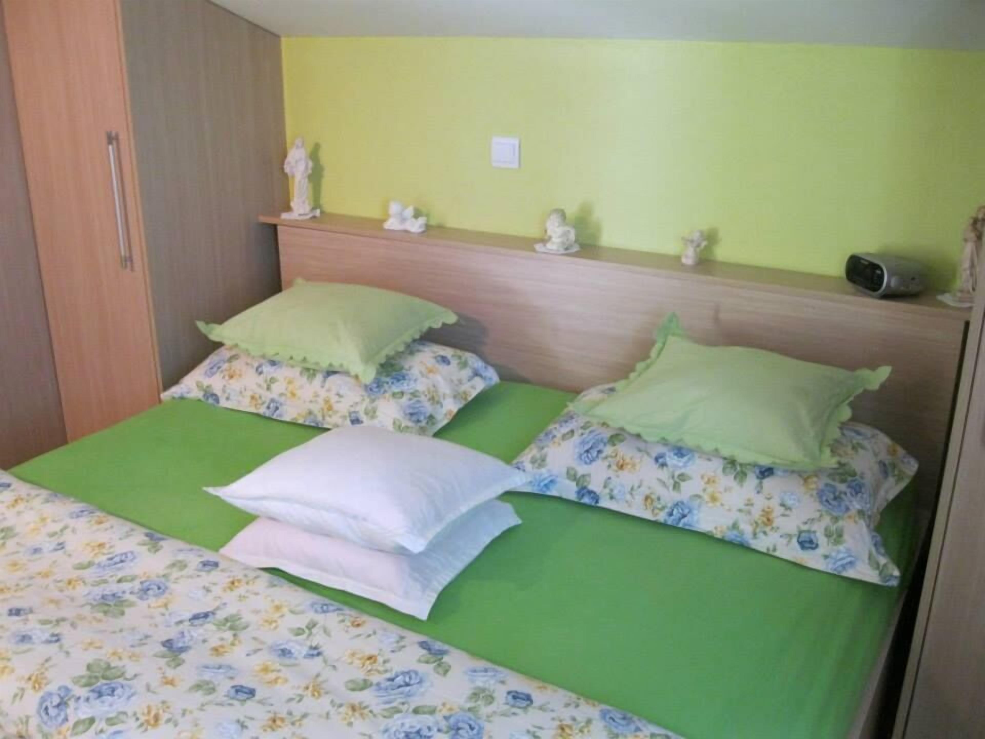 A bed with green bedding and floral-patterned pillows, a wooden headboard, and a small shelf with figurines.
